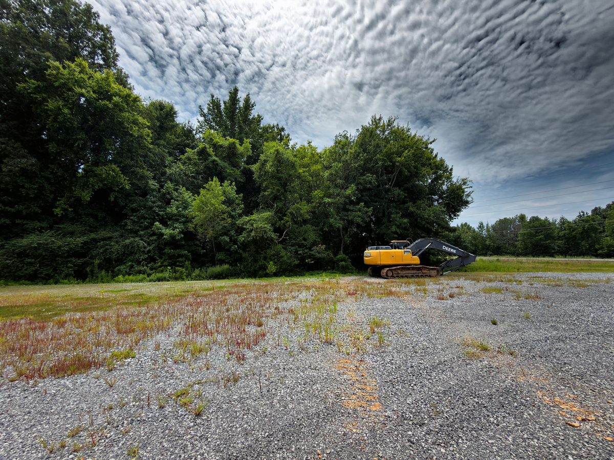 Overgrown gravel lot with standing treeline and excavator staged before land clearing in Western Kentucky